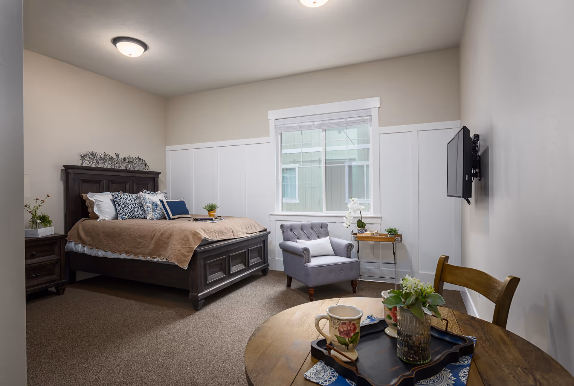 A cozy bedroom in Aspen Ridge Residences of Lehi featuring a dark wooden bed with beige bedding and decorative pillows, a matching nightstand with a small plant, a gray upholstered armchair next to a window with white blinds, a small side table with a white orchid and other plants, a wall-mounted flat-screen TV, and a round wooden table with a tray holding a floral mug and a small plant.