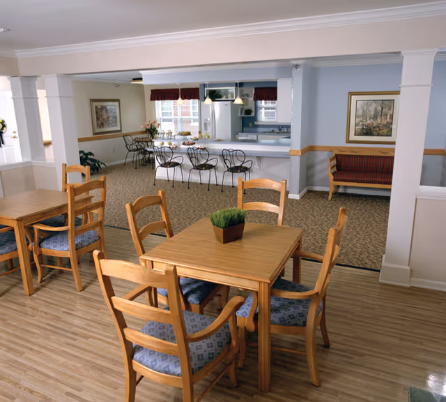A dining area in a senior living facility with wooden tables and chairs featuring blue patterned cushions. In the background, there is a kitchen counter with bar stools and pendant lights hanging above. The room has light blue walls, framed artwork, and a small bench with red upholstery along the wall.