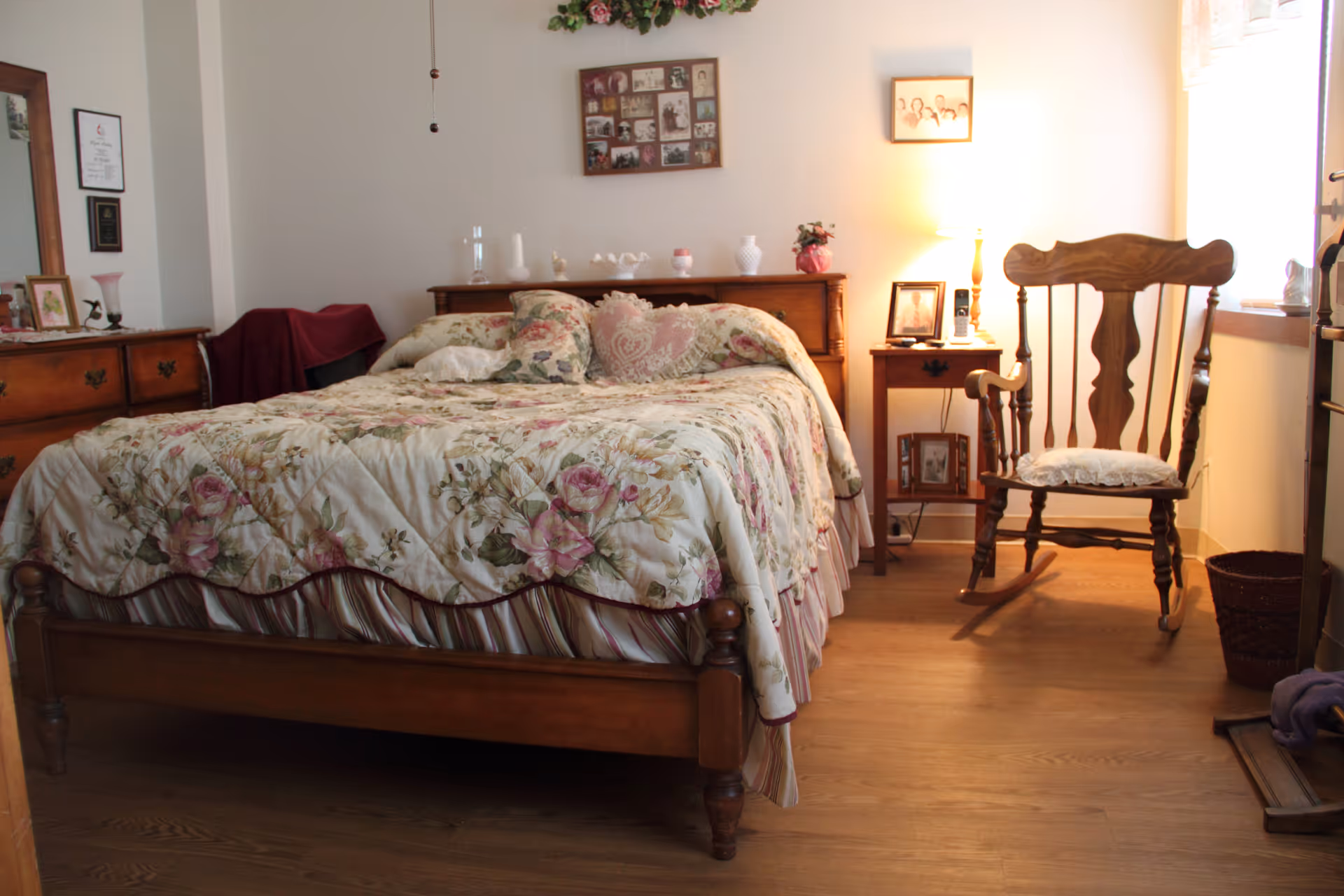 A cozy bedroom with a wooden bed frame and floral bedspread. There is a wooden rocking chair with a cushion near a window letting in natural light. A wooden dresser with framed photos and decorative items is on the left, and a small wooden nightstand with a lamp and more framed photos is on the right. The room has wooden flooring and light-colored walls with framed pictures hanging.