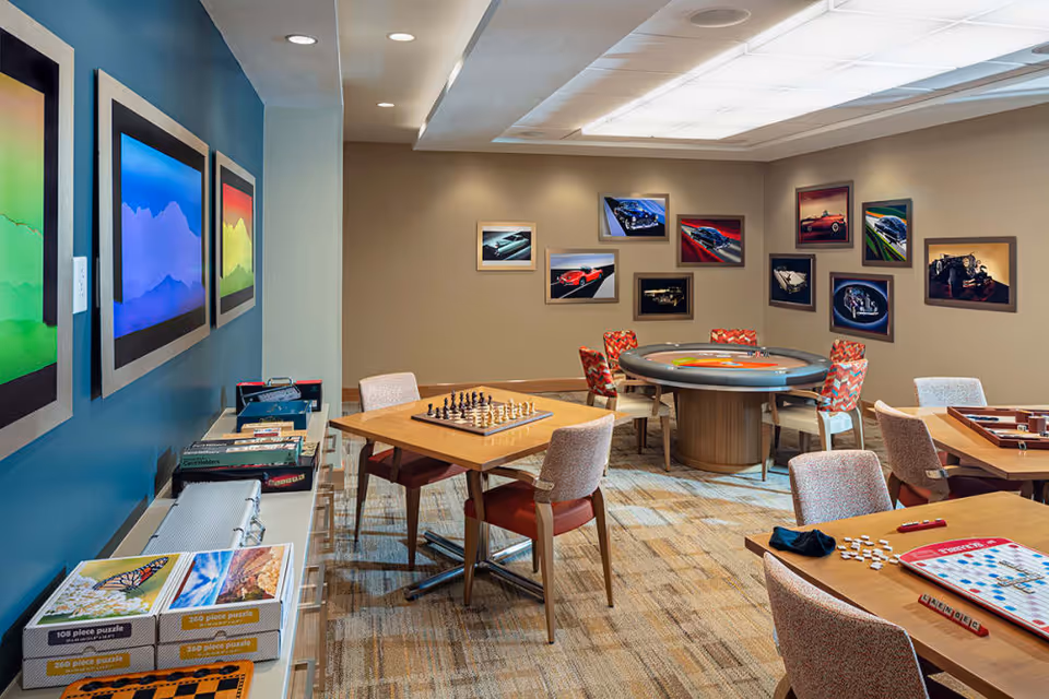 A well-lit activity room with several tables and chairs arranged for games and socializing. One table has a chessboard set up, another has a Scrabble game in progress, and a round table in the back has a roulette-style game. The walls are decorated with colorful framed pictures, including abstract art and vintage car photographs. Puzzle boxes and board games are stacked on a side shelf.