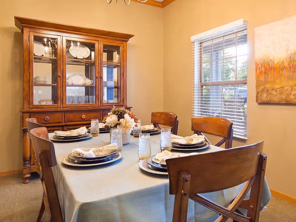 A dining room with a set table covered by a light tablecloth, wooden chairs, a floral centerpiece, and a wooden china cabinet by a window.