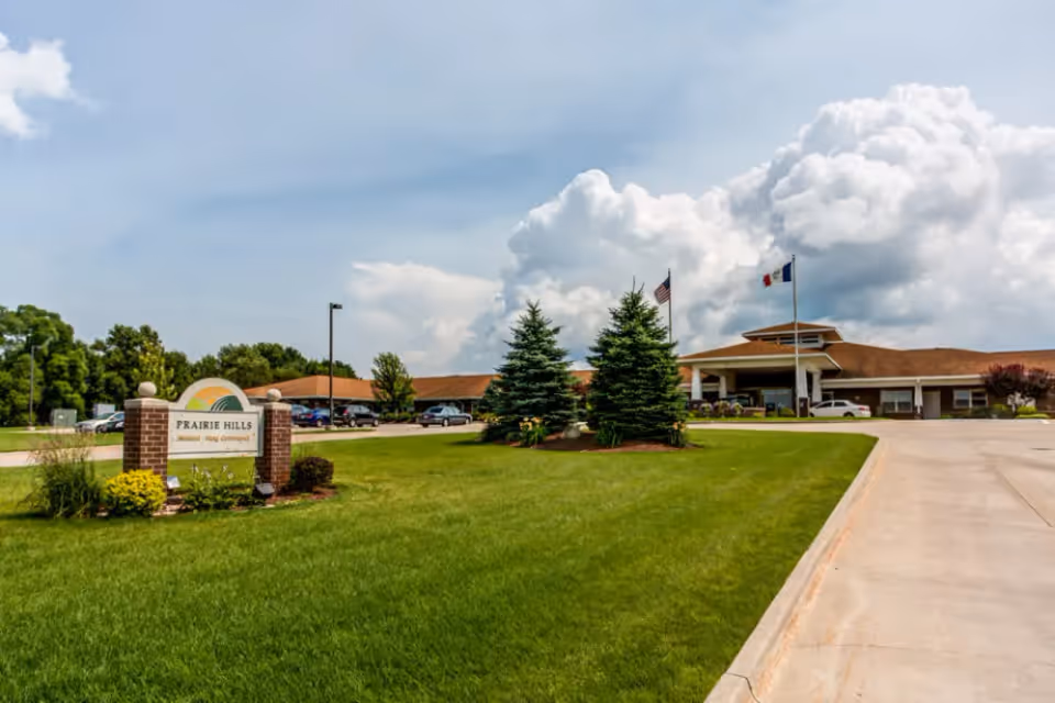 Front exterior of a single-story senior living facility with a green lawn, driveway, flagpoles, and a brick sign reading 'PRAIRIE HILLS'.