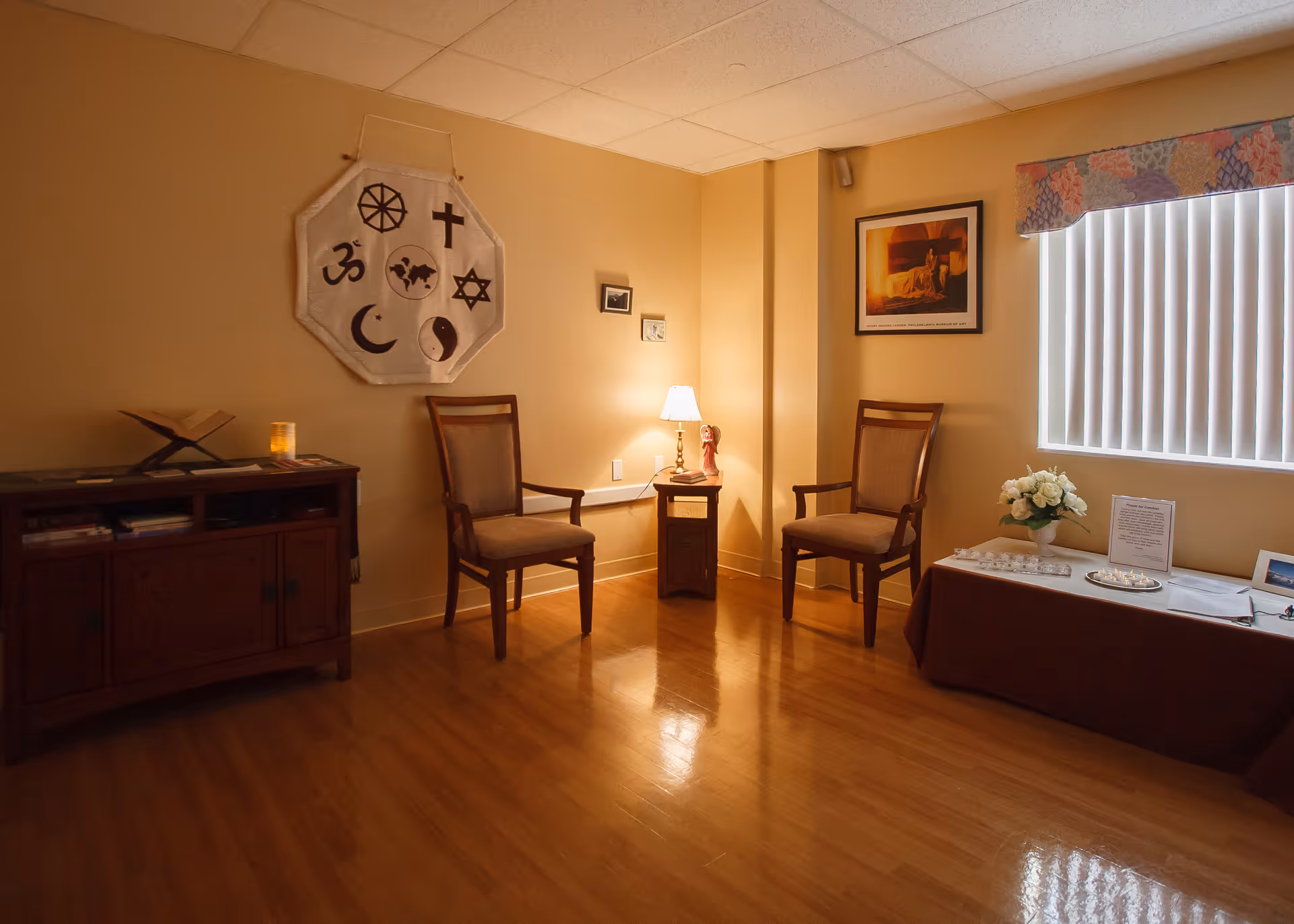 A small, warmly lit room with two wooden chairs with cushions, a small table with a lamp and a figurine, a cabinet with books and a candle, and a table covered with a brown cloth holding a vase of white flowers and some papers. The walls are beige, and there is a window with vertical blinds and a colorful valance. On the wall hangs a cloth with various religious symbols and a framed picture.