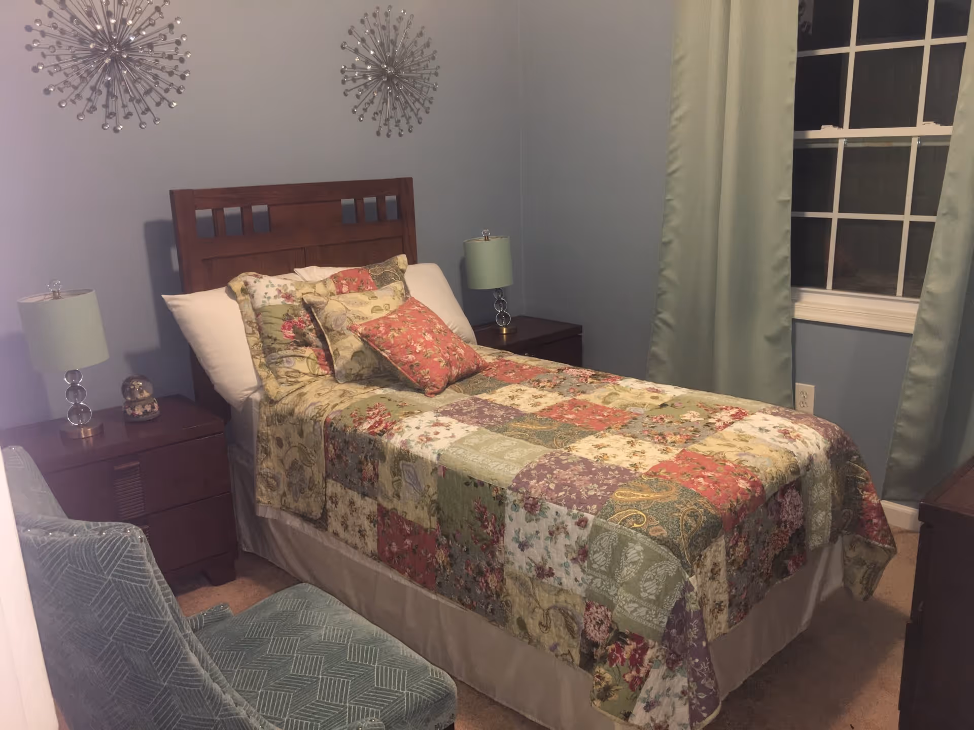 A cozy bedroom with a wooden headboard bed covered in a colorful patchwork quilt and multiple pillows. There are two matching nightstands on either side of the bed, each with a lamp. A green upholstered chair is positioned near the foot of the bed. The walls are painted light blue, and there are two decorative wall hangings above the bed. A window with green curtains is on the right side of the room.