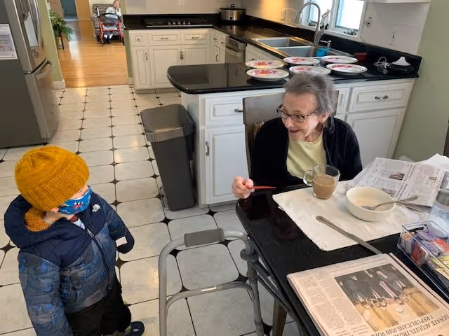 An elderly woman sitting at a kitchen table with a cup of coffee, a bowl, and newspapers in front of her, smiling and interacting with a young child wearing a yellow knit hat, blue jacket, and face mask. The kitchen has white cabinets, a black countertop, and a tiled floor. In the background, another elderly person is visible sitting in a wheelchair in a hallway.