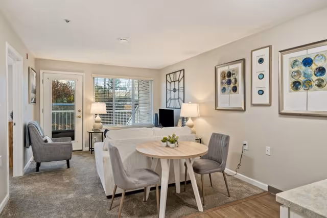 Bright living room with a sofa, round dining table and chairs, armchair, TV, lamps, and framed artwork with a balcony door and window.
