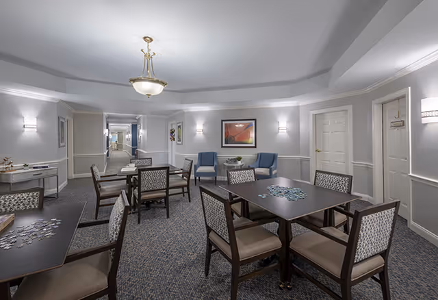 Well-lit senior living common activity/dining room with several tables and chairs, puzzle pieces on the tables, and lounge seating along the walls.