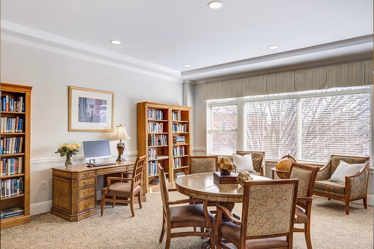 A bright and cozy common room with large windows letting in natural light. The room features a round wooden table surrounded by four upholstered chairs, two wooden bookshelves filled with books, a wooden desk with a computer and lamp, and two cushioned armchairs with pillows near the window. The carpet has a subtle pattern, and the walls are painted light with crown molding.