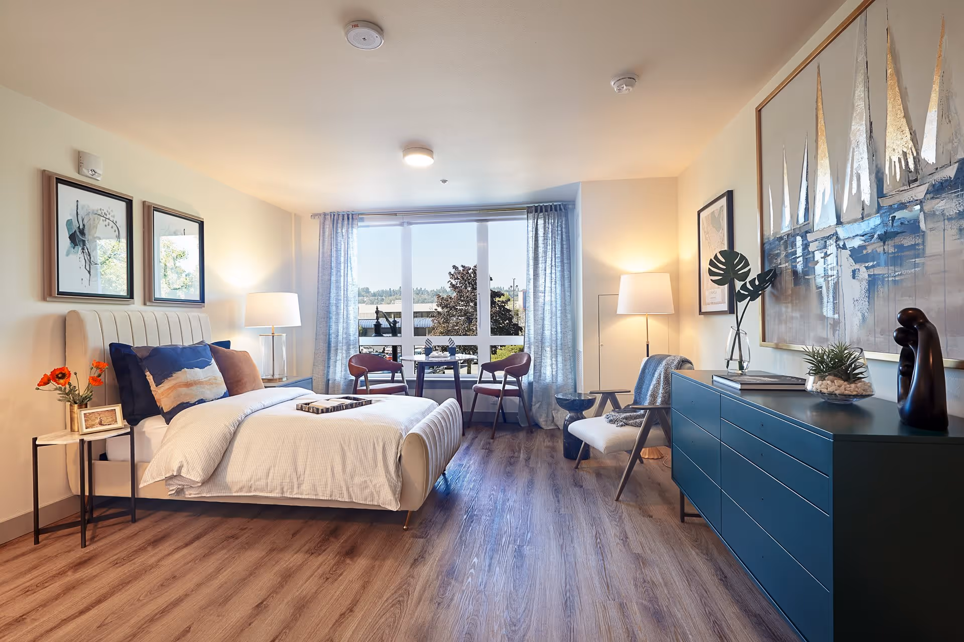 A bright and modern bedroom with a large window letting in natural light. The room features a bed with white bedding and decorative pillows, two framed artworks above the bed, a small side table with flowers and a photo frame, a blue dresser with decorative items, a comfortable chair with a throw blanket, and a small table with two chairs near the window. The floor is wood, and the walls are light-colored.
