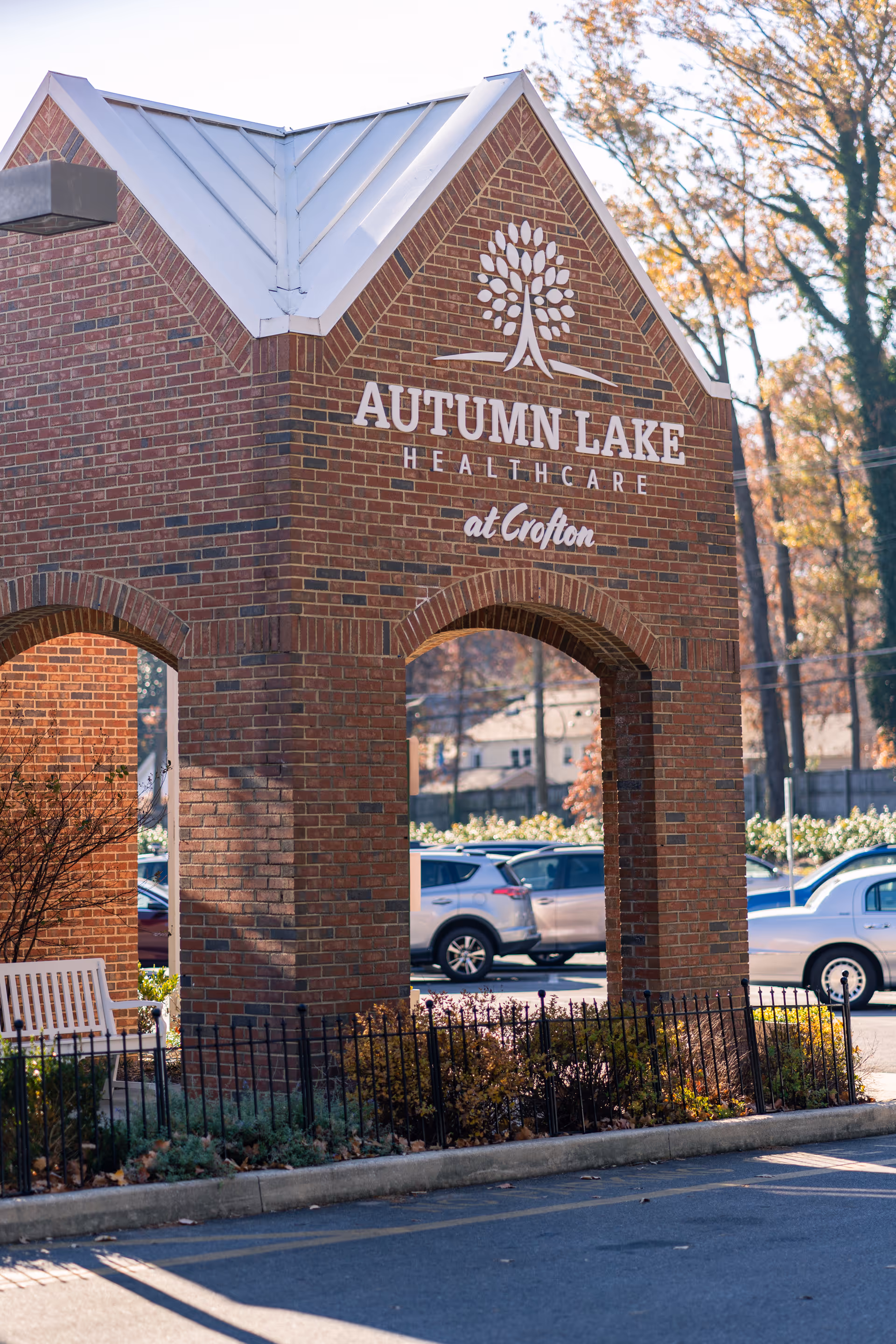 Brick arched entrance sign reading "Autumn Lake Healthcare at Crofton" with parked cars and trees in the background.