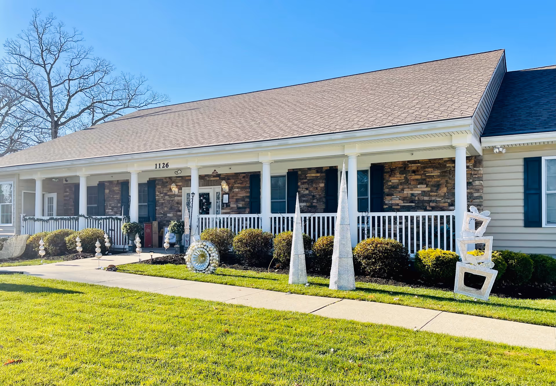 Exterior view of a single-story building with a porch supported by white columns, stone and siding facade, green shutters on windows, and decorative garden sculptures on the lawn in front. The building number 1126 is visible above the entrance door.