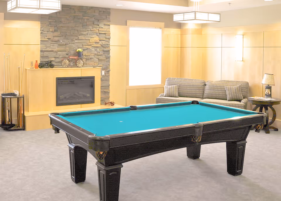 A recreational room with a pool table in the center, a stone fireplace with decorative items on the mantel, a patterned couch with cushions, a side table with a lamp, and light wood-paneled walls.