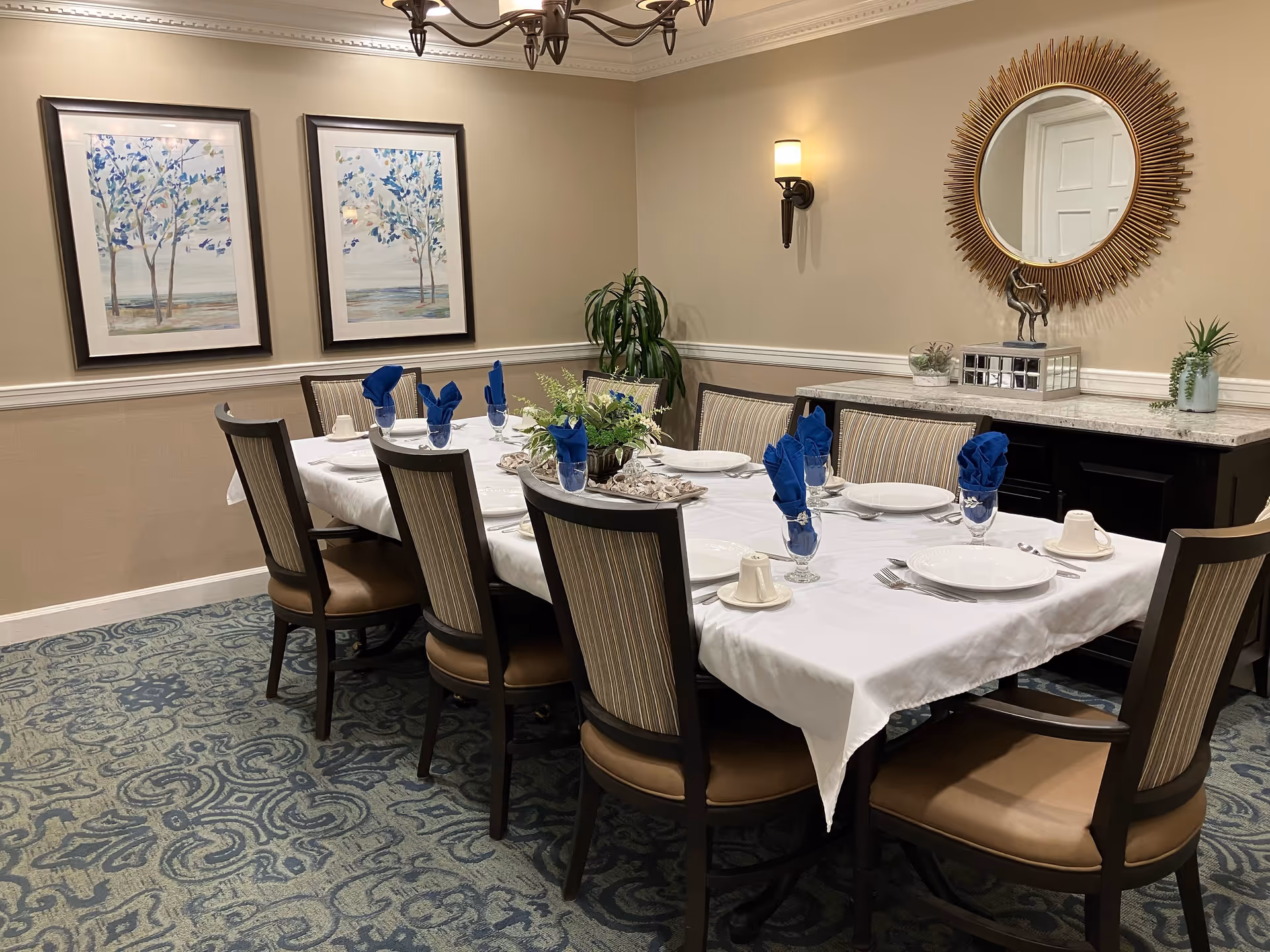 A formal dining room with a rectangular table covered with a white tablecloth, set with white plates, silverware, glassware with blue folded napkins, and small cups. The room has eight chairs with striped upholstery, two framed paintings of trees on the wall, a decorative round mirror above a sideboard with plants and decor, and a patterned carpet.