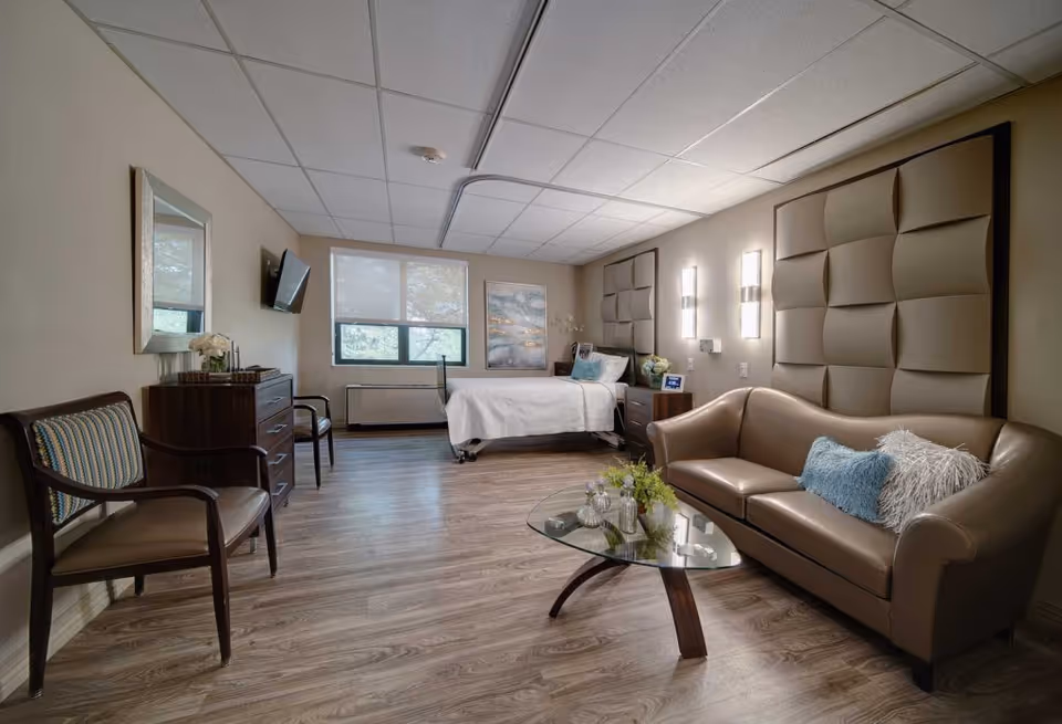 A spacious, well-lit bedroom in a senior living facility featuring a single bed with white bedding, a brown leather sofa with decorative pillows, a glass coffee table with small plants and decor, two wooden chairs with cushions, a dresser with a mirror above it, a wall-mounted TV, and large windows with blinds allowing natural light to enter.