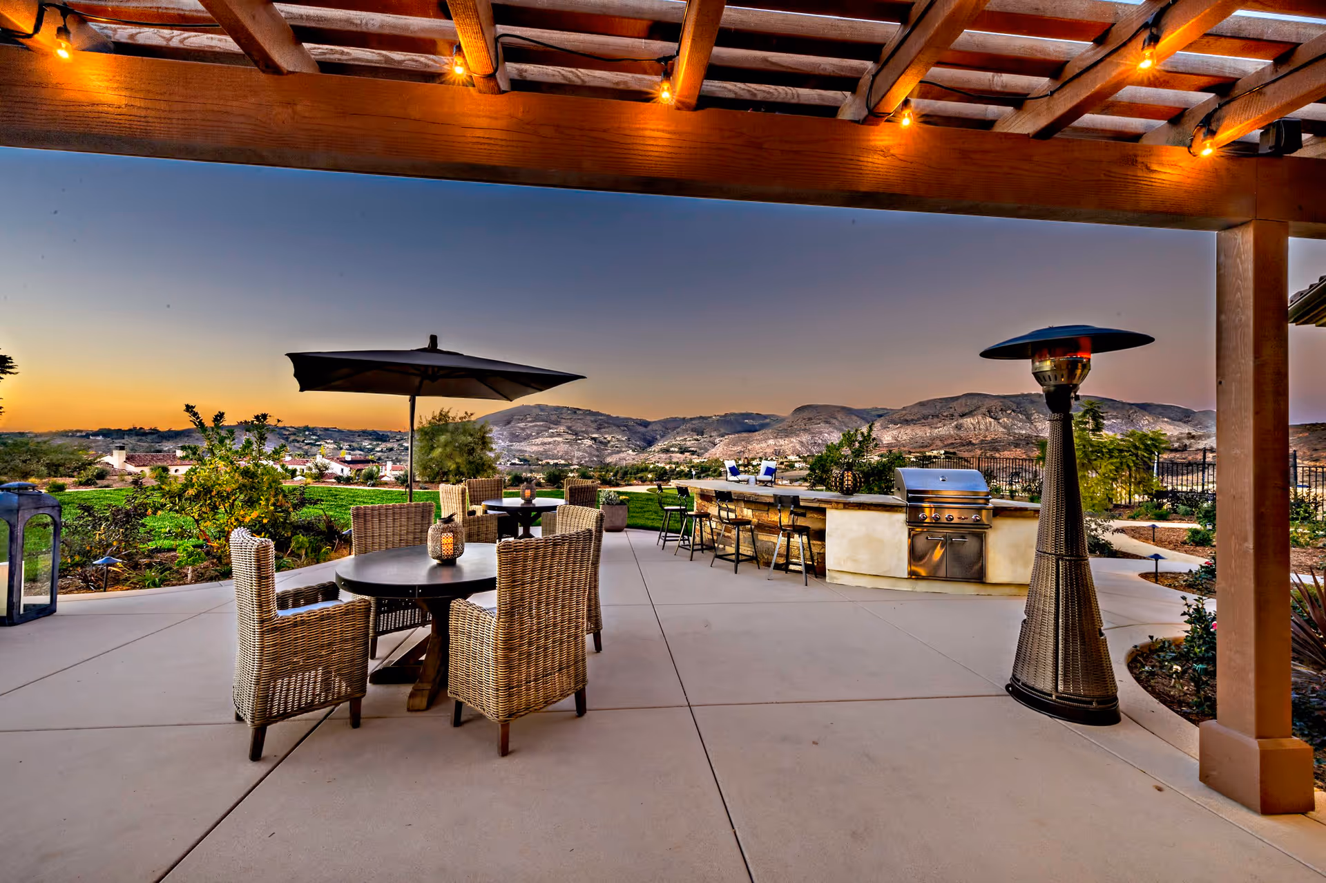 Outdoor patio area at sunset with wicker chairs around a round table under a large umbrella, a tall outdoor heater, and a built-in grill with bar seating. The patio overlooks a scenic view of hills and greenery under a clear sky.