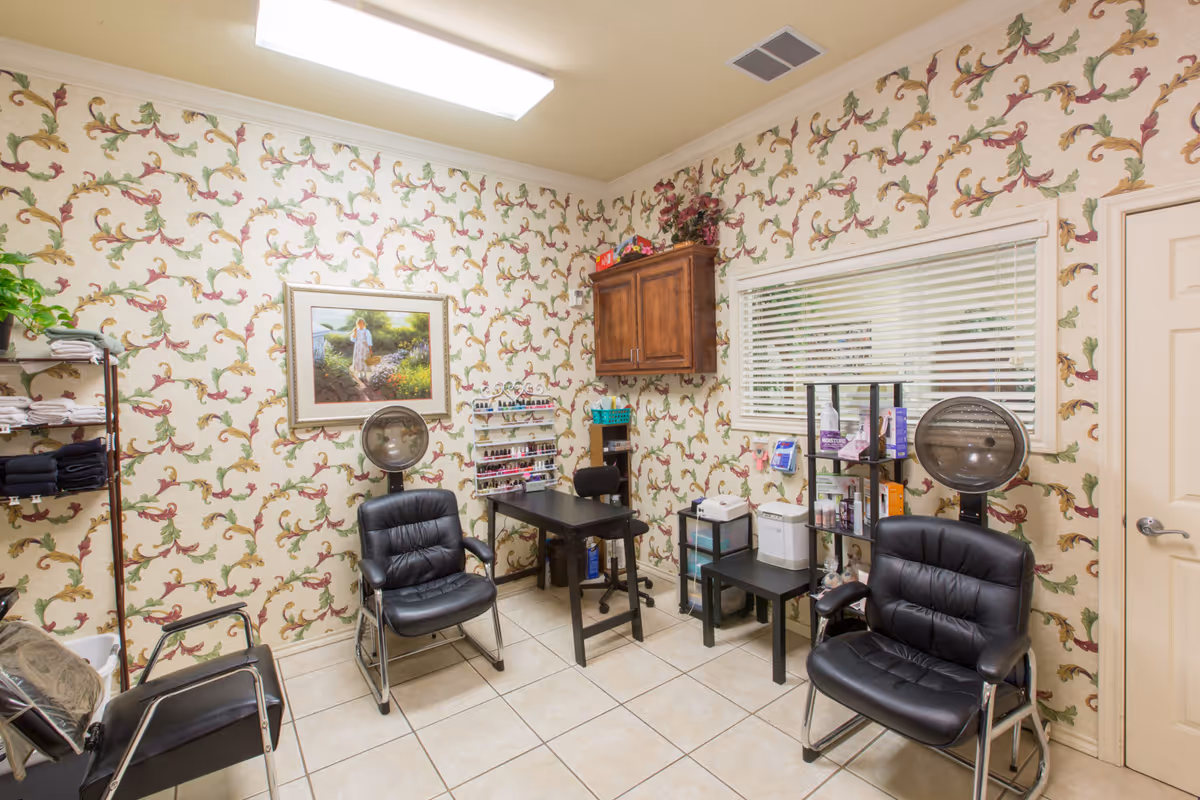 Small salon-style room with black leather chairs, hooded hair dryers, nail polish racks and floral wallpaper.