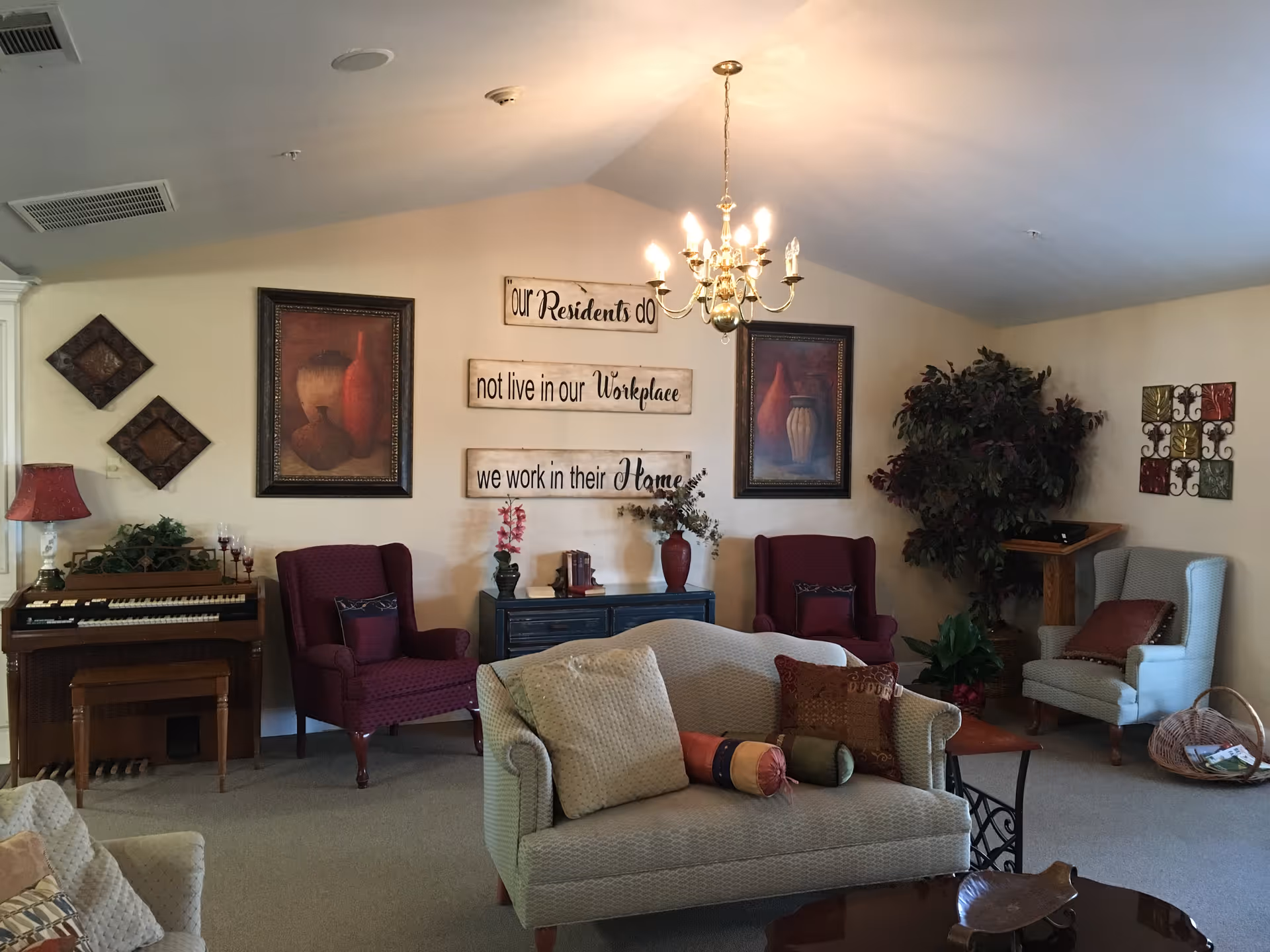 A cozy living room area in a retirement facility with a beige sofa adorned with multiple pillows, two burgundy armchairs, and a light blue armchair. The walls are decorated with framed artwork and a three-part wooden sign that reads 'Our Residents do not live in our Workplace we work in their Home.' A chandelier hangs from the ceiling, and there is a piano with a red lamp on the left side. There are also plants and a small wooden podium in the corner.