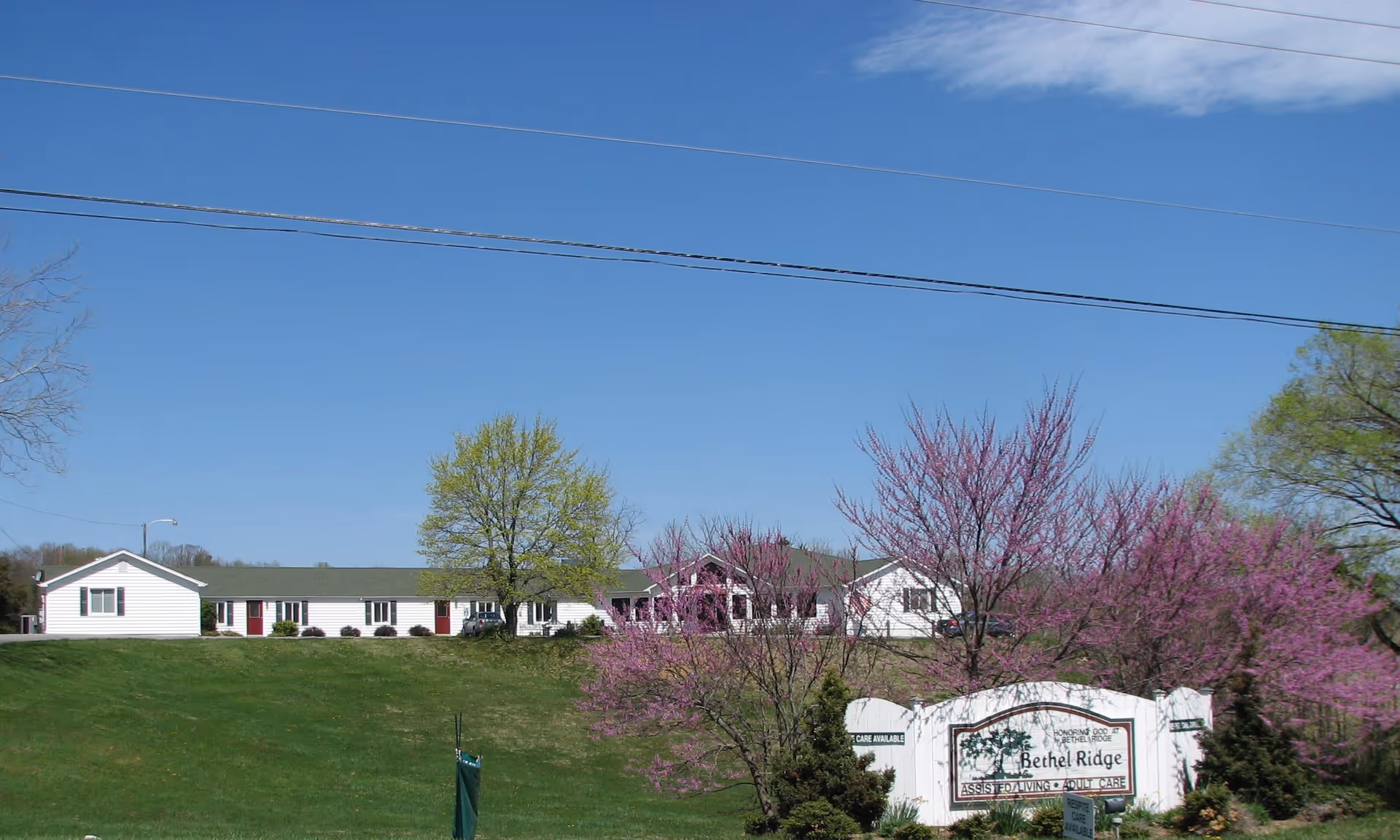 Exterior view of Bethel Ridge Assisted Living facility showing a single-story white building with a green roof, surrounded by green grass, trees with green and purple foliage, and a clear blue sky. A white sign with the facility's name is visible in the foreground.