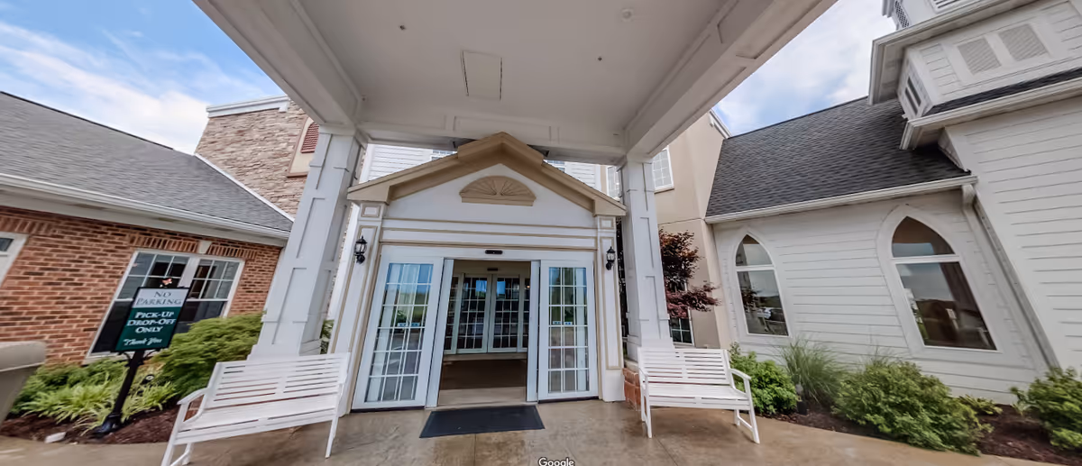 Entrance of Doverwood Skilled Nursing Facility showing a covered drop-off area with two white benches on either side of automatic sliding glass doors. The building exterior features a combination of brick and white siding with decorative windows and landscaping around the entrance.