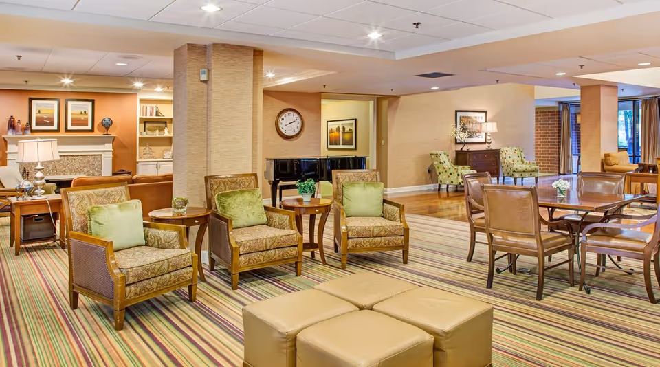 Spacious senior living common room with upholstered armchairs, dining tables, a piano, fireplace, and striped carpet.