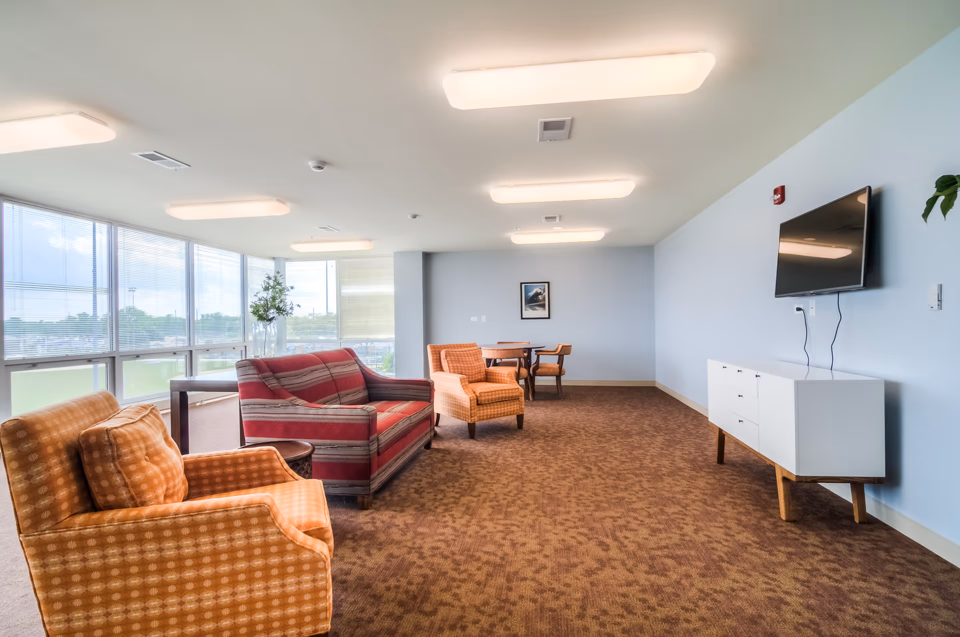 A spacious senior living facility common area with large windows letting in natural light. The room features a red striped sofa, two orange patterned armchairs, a small round table, a white media console with a wall-mounted flat-screen TV, and a small dining table with four chairs. The walls are painted light blue and the floor is covered with brown carpet.