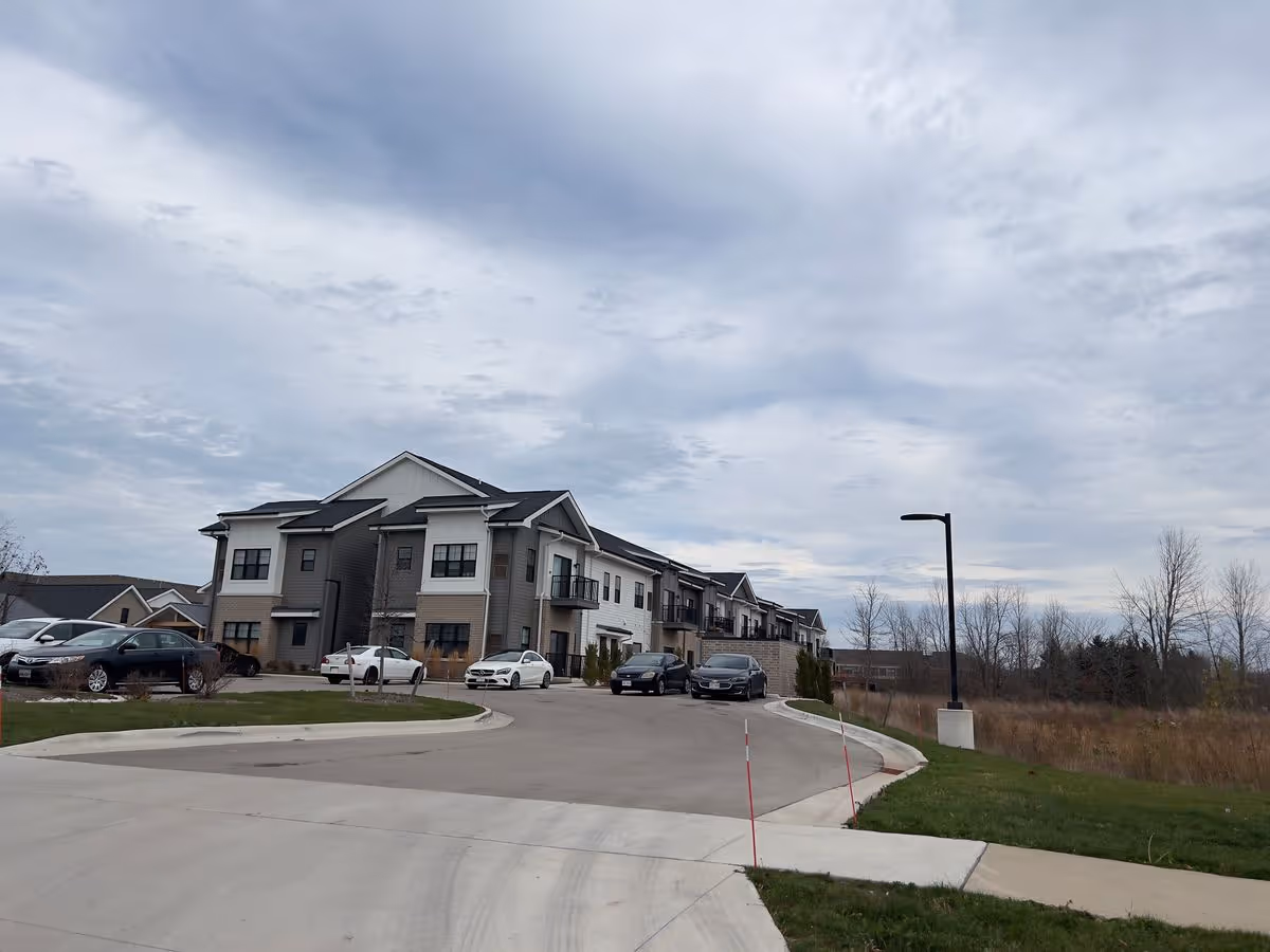 Exterior view of a two-story assisted living building with a curved driveway, parked cars, and a cloudy sky.