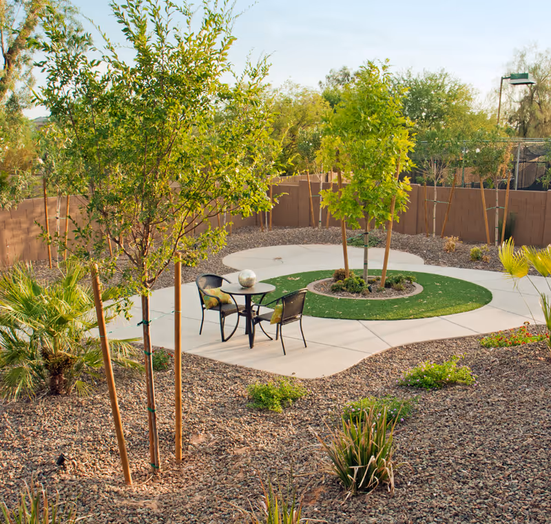 Small landscaped outdoor courtyard with a circular paved path, a table and two chairs, young trees, and gravel groundcover.