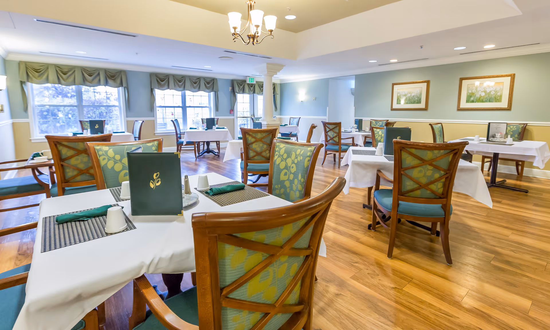 A bright and spacious dining room with wooden floors, several tables covered with white tablecloths, green patterned chairs, and menus placed on the tables. Large windows with green valances allow natural light to fill the room. The walls are painted light green with white trim, and two framed floral paintings hang on one wall. A chandelier hangs from the ceiling.