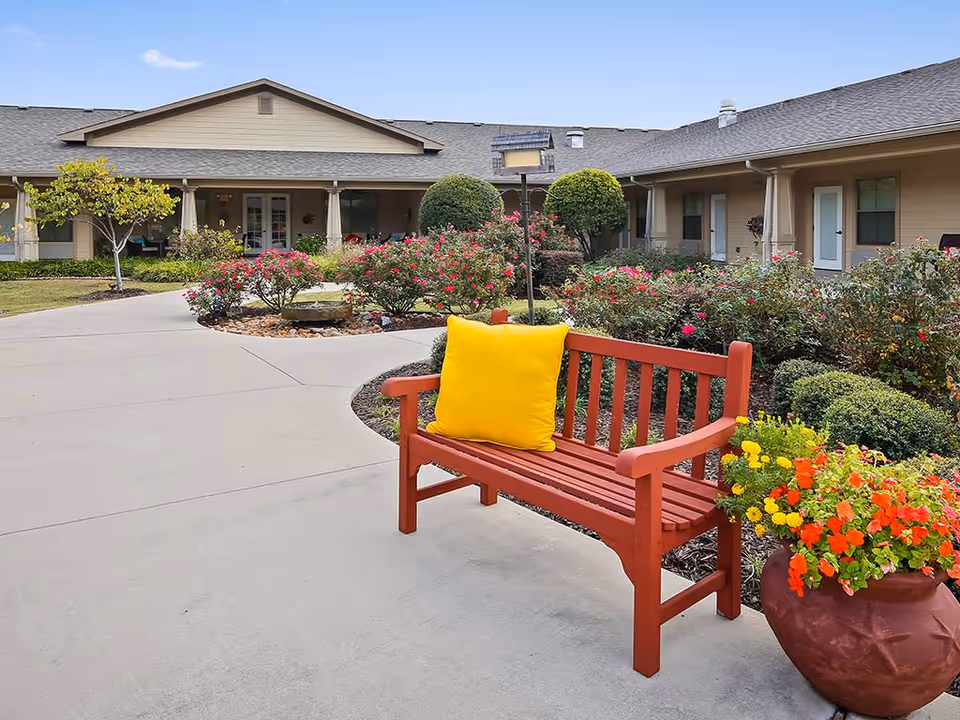 Red wooden bench with a yellow pillow in a landscaped courtyard surrounded by flowers and a single-story assisted living building in the background.