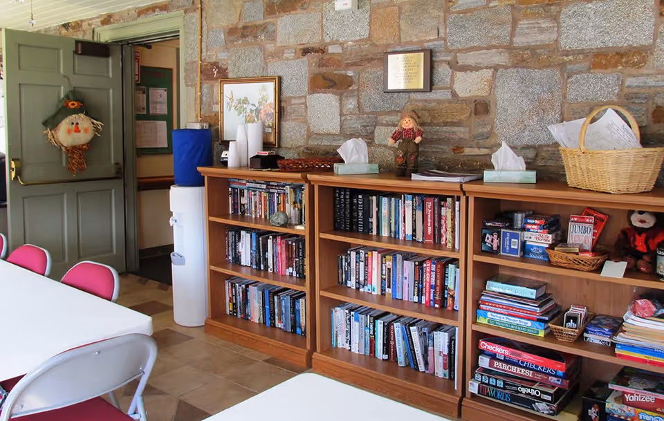 Interior room with stone walls featuring wooden bookshelves filled with books, board games, and decorative items. There are tables and chairs with red seats in the foreground, a water cooler near an open door with a scarecrow decoration, and framed artwork on the wall.