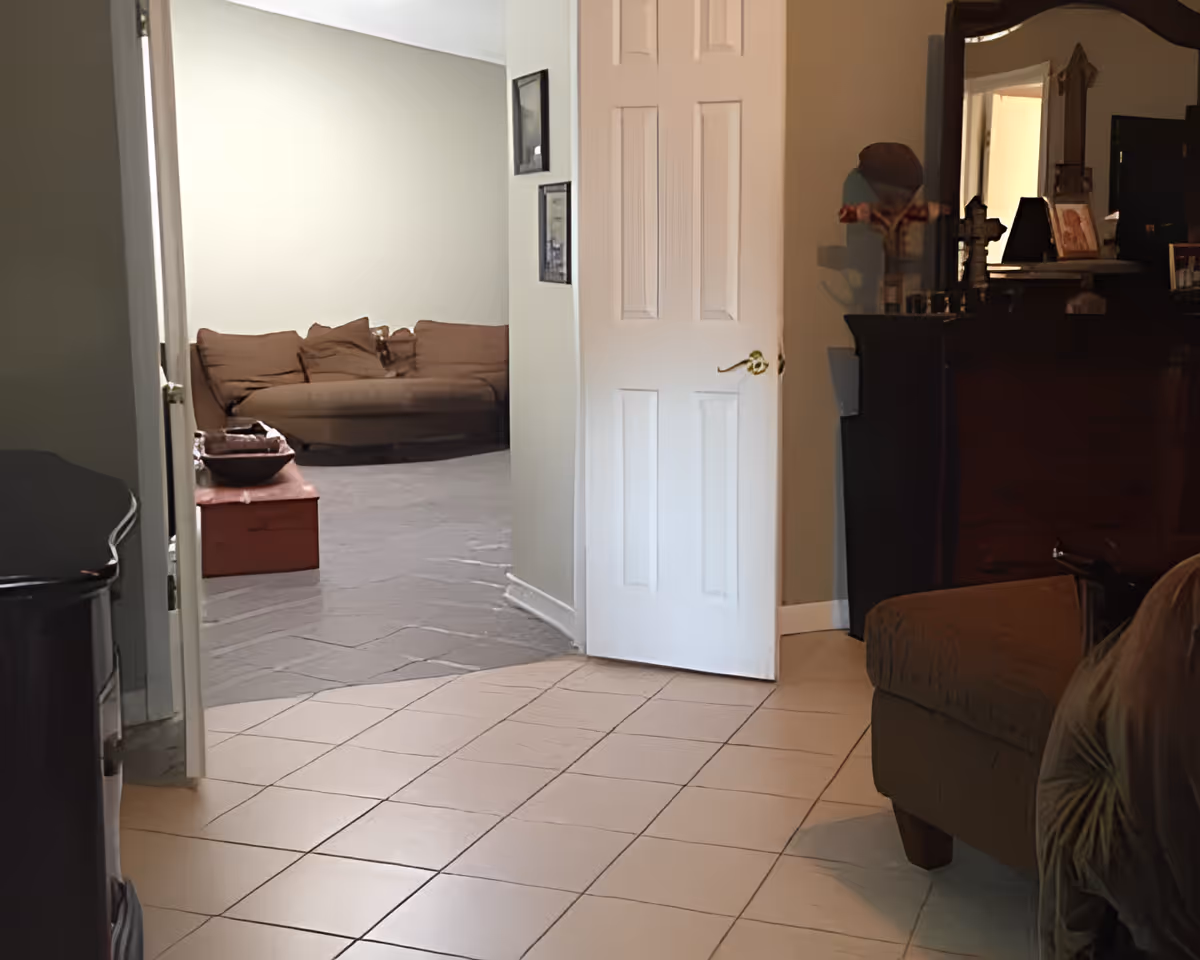 View of an interior living space with a tiled floor in the foreground and a carpeted area with a brown sofa in the background. There is a white door partially open, a wooden chest, and a dark wooden dresser with a mirror and decorative items on top.