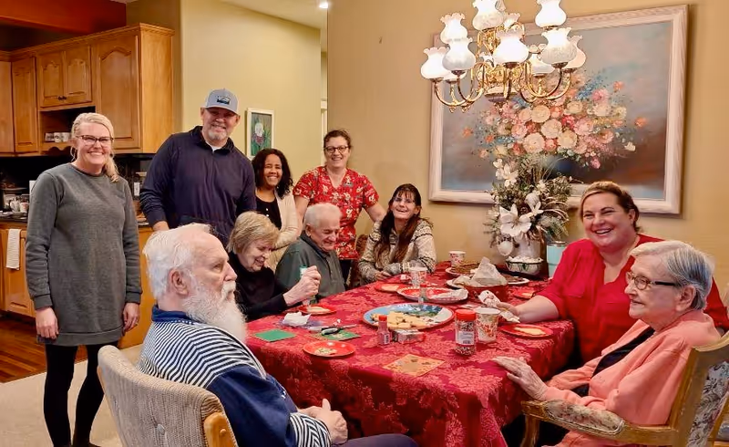 A group of elderly and middle-aged people gathered around a dining table covered with a red tablecloth. The table has plates, cups, and snacks. The setting is a cozy dining room with a chandelier overhead and a large floral painting on the wall. Some people are seated while others stand behind them, all smiling and engaging with each other.