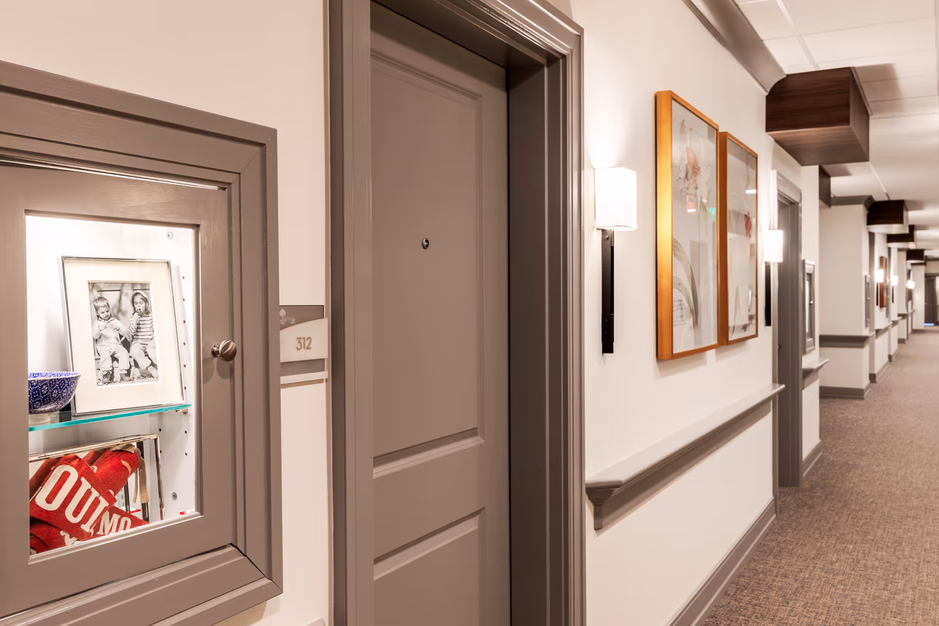 A well-lit hallway in a senior living facility with beige walls and carpeted floor. The hallway features closed gray doors, wall-mounted light fixtures, framed artwork, and a small display cabinet with a black and white photo and decorative items. The door number 312 is visible.