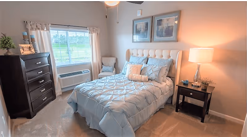 A cozy bedroom with a neatly made bed featuring a tufted headboard and light blue bedding. There is a dark wooden dresser with decorative items on top to the left, a window with white curtains behind it, and a small upholstered chair in the corner. On the right side of the bed, there is a dark wooden nightstand with a lamp and small decorative plants. Two framed pictures hang on the wall above the bed.