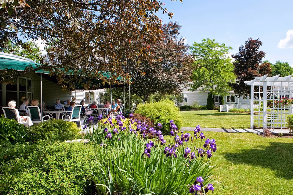 A group of elderly people sitting and socializing under a green awning in a garden area with lush greenery, purple flowers, trees, and a white pergola on a sunny day.