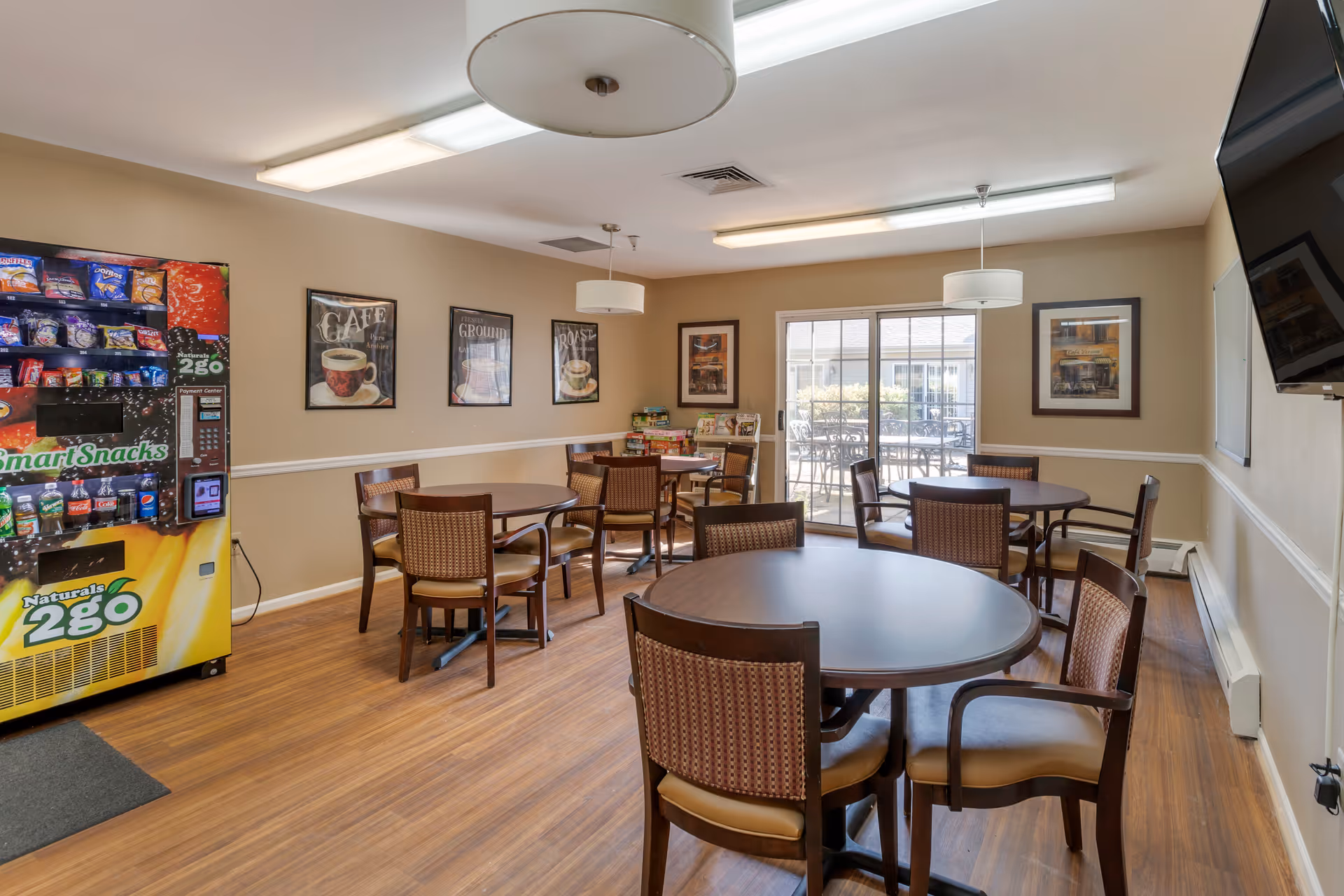 A well-lit dining area with round tables and chairs arranged neatly. There is a vending machine stocked with snacks and drinks on the left side. The walls are decorated with framed pictures, and large windows at the back allow natural light to enter, showing an outdoor patio area with tables and chairs. Ceiling lights hang above the tables, and a television is mounted on the right wall.