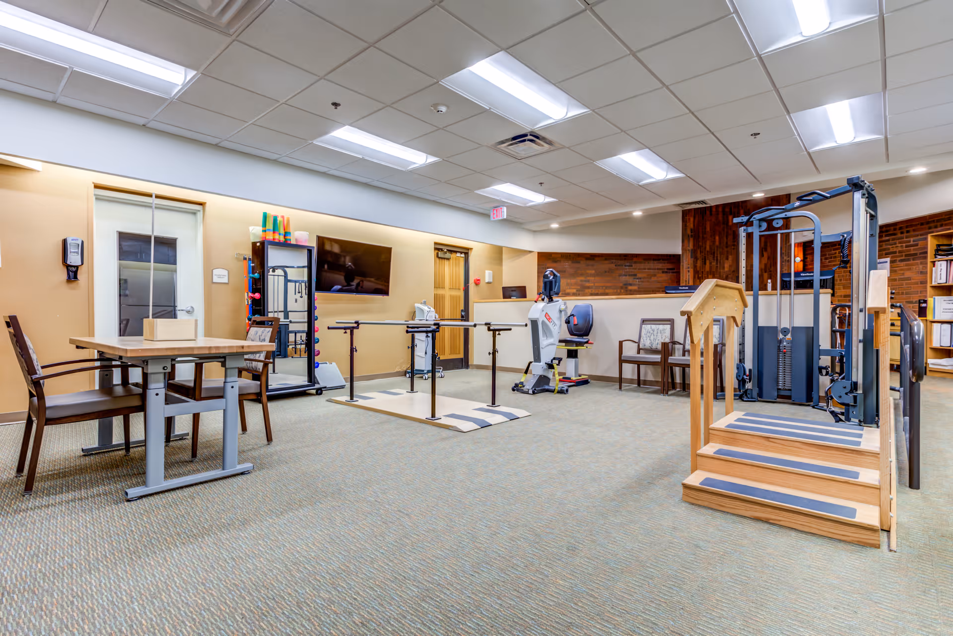 A physical rehabilitation room with exercise equipment including parallel bars, a stationary exercise bike, a multi-functional weight machine, and a wooden staircase with handrails for therapy. There is a table with two chairs on the left side, a wall-mounted TV, and shelves with colorful exercise tools. The room has a carpeted floor, beige walls, and a drop ceiling with fluorescent lighting.