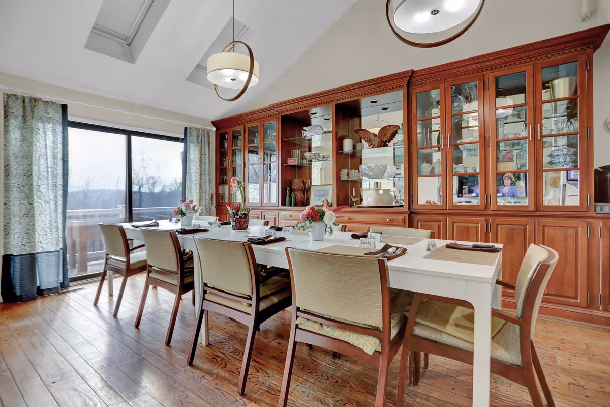 A dining room with a long white table set with placemats, napkins, and name cards. There are eight chairs around the table. Behind the table is a large wooden cabinet with glass doors displaying various decorative items and glassware. Large windows with patterned curtains allow natural light to fill the room, and two modern pendant lights hang from the ceiling.
