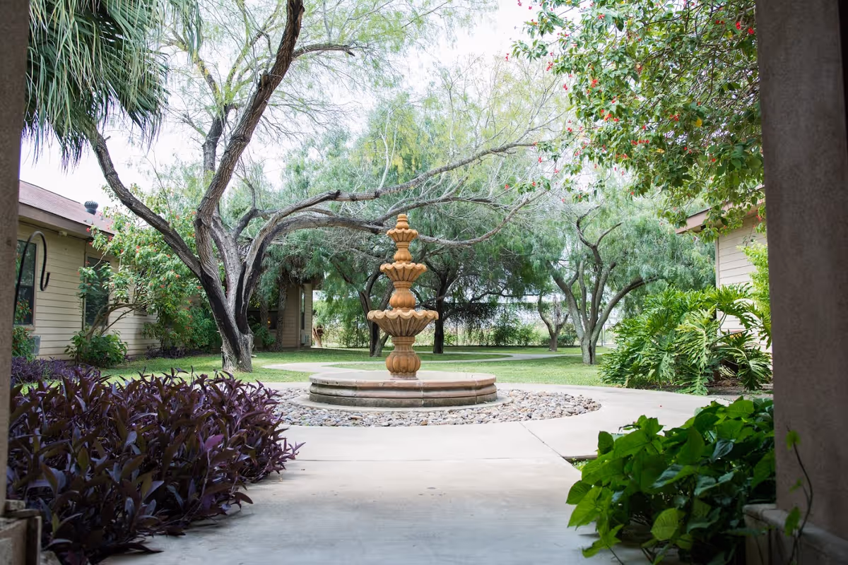 A peaceful outdoor courtyard featuring a multi-tiered stone fountain surrounded by a circular stone border and landscaping. The area is framed by trees, bushes, and plants, with pathways leading through the garden space between buildings.