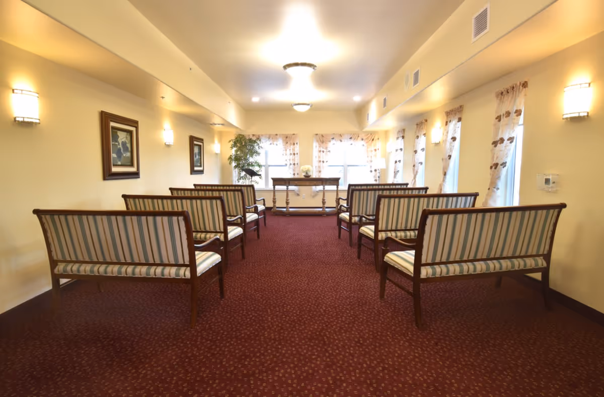 A small meeting or chapel room with six wooden benches upholstered in striped fabric arranged in two rows facing a wooden table with a flower arrangement. The room has beige walls, red carpet, framed artwork, wall sconces, and windows with sheer floral curtains letting in natural light.