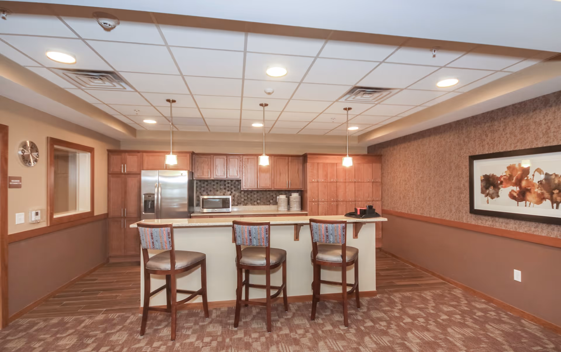 Interior view of a senior living facility kitchen area with a countertop bar and three high chairs. The kitchen features wooden cabinets, a stainless steel refrigerator, a microwave, and pendant lights hanging from the ceiling. There is a patterned carpet on the floor and a framed abstract artwork on the right wall.