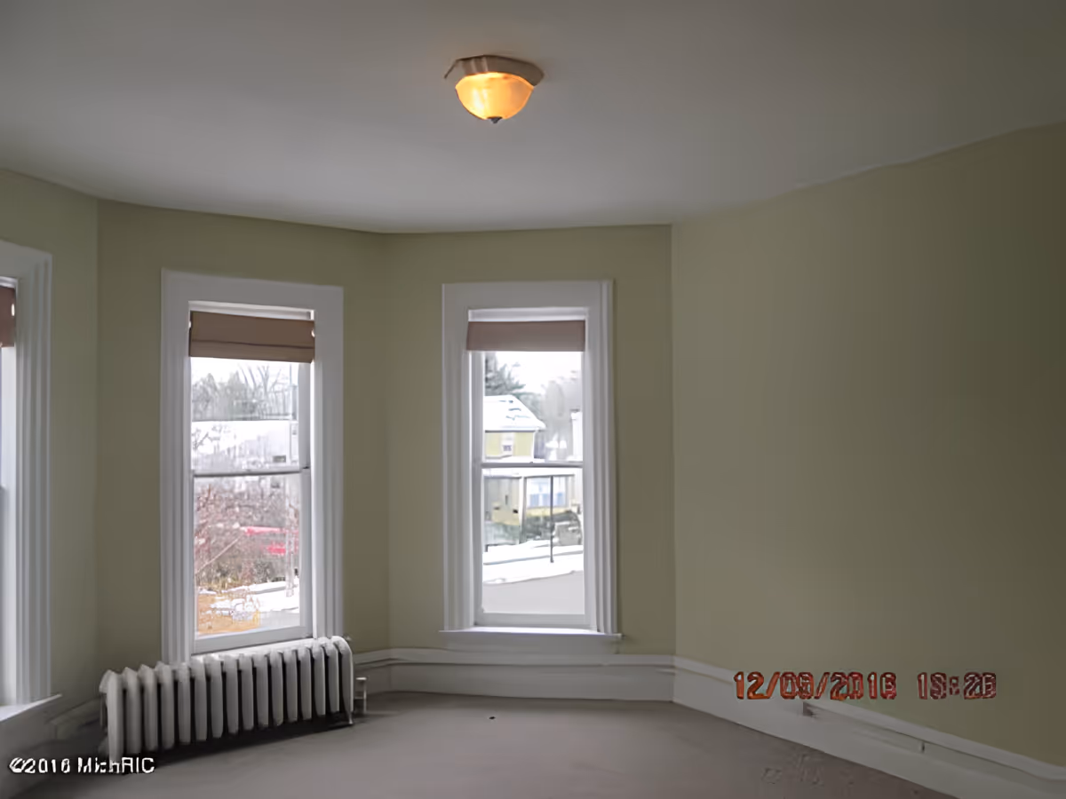 Empty room with three tall bay windows, pale green walls, a radiator under one window, and a ceiling light.