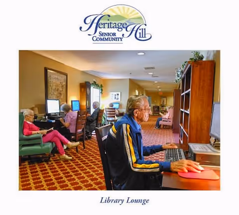 Several elderly individuals seated at computer stations in a cozy library lounge with patterned carpet and warm lighting at Heritage Hill Senior Community.