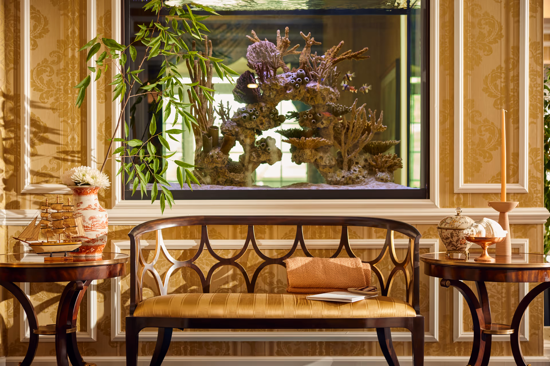 A cozy seating area with a wooden bench featuring a cushioned seat and a folded blanket with a book on it. On either side of the bench are round wooden side tables, one holding a decorative ship model and a vase with flowers, the other holding a candle and a decorative jar. Behind the bench is a large aquarium with coral and small fish, set against a wall with ornate gold-patterned wallpaper and white panel molding.