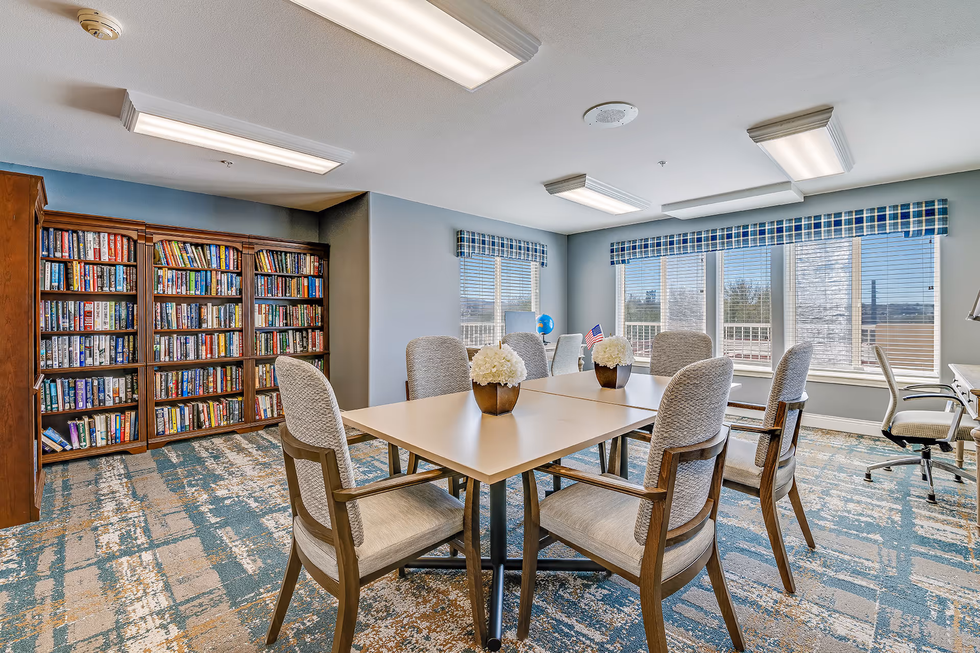 A bright room with a large table surrounded by eight cushioned chairs. Two small flower arrangements are placed on the table. The room has large windows with blue plaid valances letting in natural light. A wooden bookshelf filled with books is against one wall, and there are additional chairs and a desk near the windows. The carpet has a blue and beige abstract pattern, and the ceiling has several rectangular fluorescent light fixtures.