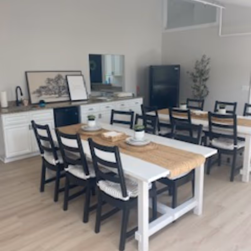 Communal dining area with two long white tables, black chairs, place settings and a kitchenette with a refrigerator in the background.