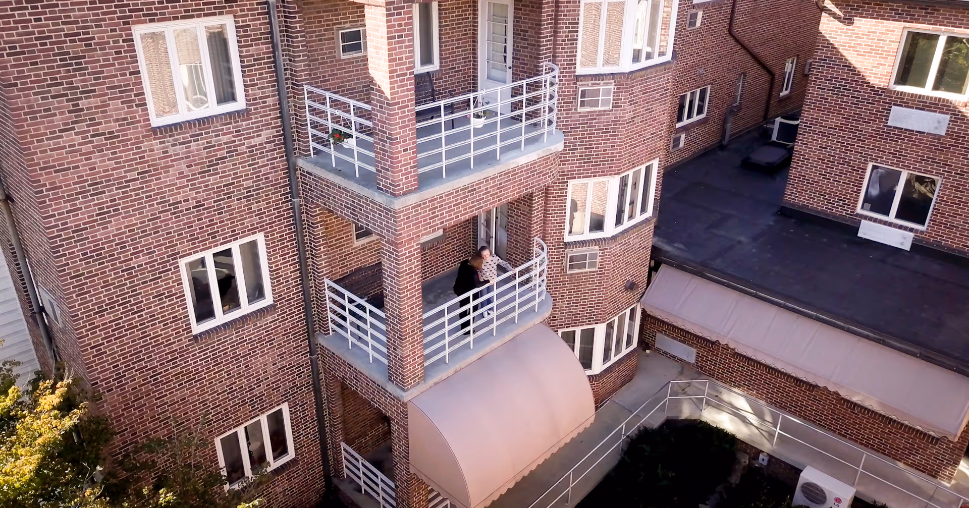 View of a brick apartment building with multiple balconies. Two people are standing on one of the balconies, looking out. The building has white railings and several windows, with a small awning over an entrance below.