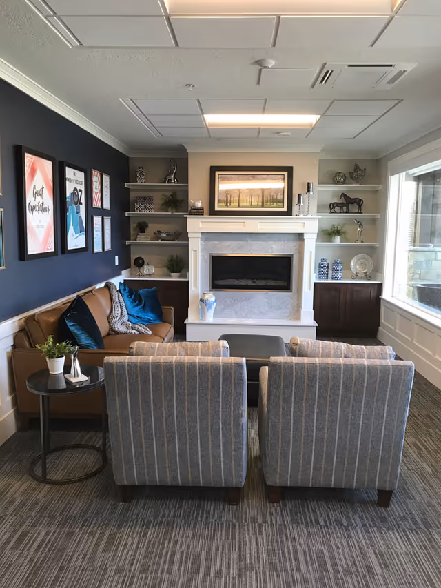 A cozy living room with a brown leather sofa adorned with blue and gray pillows, two striped armchairs facing a modern fireplace with a marble surround. The fireplace is flanked by built-in shelves decorated with plants, vases, and sculptures. A large window on the right lets in natural light, and framed artwork hangs on the dark blue wall to the left.