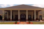 Front exterior view of a single-story building with a covered entrance supported by columns, a central doorway with glass panels, and a brick walkway leading up to the entrance. The building has a pitched roof and small landscaped areas with plants in front.