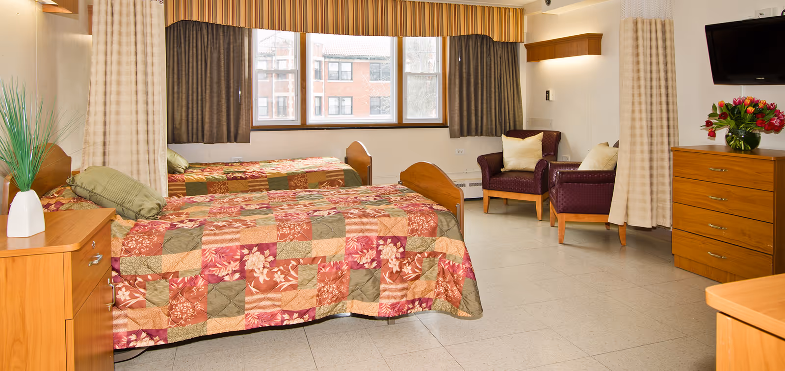 A bright and clean bedroom in a senior living facility with two twin beds covered in patchwork quilts in shades of red, green, and beige. The room has large windows with striped valances and curtains, two purple armchairs with cream pillows, a wooden dresser with a vase of red flowers, and a wall-mounted TV.